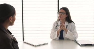 Beyaz üniformalı gülümseyen kadın doktor muayene odasında oturuyor, kadına el sıkışıyor, güvenini dile getiriyor, özel klinikte başarılı tıbbi muayeneler yapıyor.