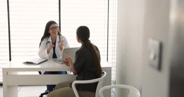 Beyaz önlüklü genç bayan doktor hastayla konuşuyor, tıbbi tavsiyeler veriyor, muayene odasındaki masada oturan sağlığın tedavi planını sunuyor. Profesyonel sağlık danışmanlığı