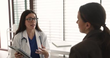 Genç pratisyen hekim elinde pano tutarken kadın hastayla konuşuyor, tedavi planını, ilave sağlık testlerini ve prosedürlerini açıklıyor, profesyonel tıbbi tavsiyeler veriyor.