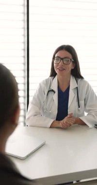 Beyaz önlüklü gülümseyen kadın doktor muayene odasında oturuyor, hastalarla tokalaşıyor arkadaşça tavırlar sergiliyor, klinikteki randevuyu başarıyla başlatıyor veya sonuçlandırıyor.