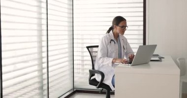 Kürklü kadın doktor, tıp bürosunda modern dizüstü bilgisayarla çalışıyor, idari ya da hastalarla ilgili görevlere katılıyor, tıbbi kayıtları inceliyor ya da muayeneden sonra notlar yazıyor.