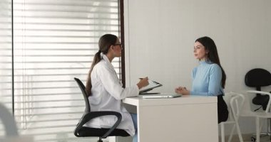 Modern muayene odasında üniformalı genç bayan doktor masa başında elinde not panosu ve kalemle dikkatle hasta kadına hastalık belirtilerini, sağlık sorunlarını, tıbbi geçmişi ve mevcut ilaçları soruyor.