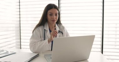 Dizüstü bilgisayarda çalışan çekici kadın doktor iş yerinde oturur, hasta tıbbi kayıtlarını inceler, laboratuvar sonuçlarını ya da görüntüleme raporlarını kontrol eder, tedavi seçeneklerini düşünür, uzaktan reçete yazar.