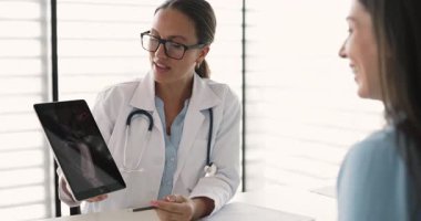 Beyaz önlüklü travmatolog tablet tutuyor ve röntgen görüntülerini ekranda gösteriyor, röntgen sonuçlarını açıklıyor, teşhis, yaralanma ya da tedavi planını kadın hastaya anlatıyor. Sağlık hizmetleri
