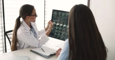 Kadın doktor beyin MR 'ını tutarken ve analiz ederken hastaya sonuçları açıklarken, taramadaki belirli alanlara işaret ederken, tıbbi bulguları veya nörolojik sağlıkla ilgili potansiyel sorunları tartışırken,