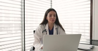 Kadın pratisyen hekimin hasta notlarını konsültasyondan sonra güncellemesi, klinikte dizüstü bilgisayarda çalışması, e-postalarına cevap vermesi, hastalarla teletıp platformu üzerinden iletişim kurması.