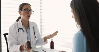 Kadın doktor masada oturuyor, elinde haplarla hastayla konuşuyor, ilaçların amacını, dozunu ya da talimatlarını anlatıyor. Reçete, tedavi, tıbbi danışma