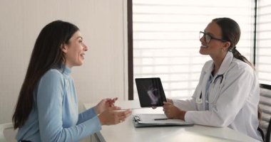 Paltodaki kadın doktor, eklem röntgenini dijital tablette gösteriyor, tarama sonuçlarını açıklıyor, olası yaralanmaları, iltihaplanmayı veya sorunu tartışıyor, sağlık durumu ve tedavi seçenekleri sunuyor