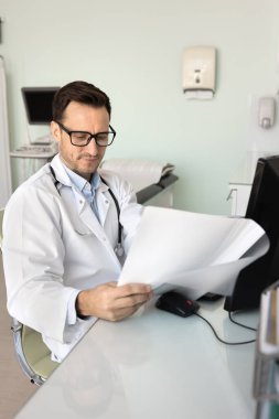 Gözlüklü ve beyaz önlüklü doktor işyerindeki tıbbi belgeleri dikkatle inceliyor, evrak işlerini incelerken odaklanmış görünüyor, hasta kayıtlarını, test sonuçlarını veya tedavi planlarını incelerken