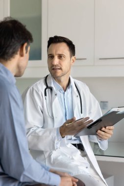 Klinik hastasıyla tartışan, pano ve kalem tutan, teşhisi açıklayan, sağlık şikayeti yazan, hastalık belirtilerini soran, tıbbi danışmanlık sırasında sağlık hizmetleri tavsiyesi veren ceketli doktor.