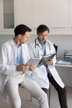 İki Latin doktor pano tutuyor, not alıyor, dijital tablet kullanıyor, profesyonel konuşmalar yapıyor, tıbbi verileri ve hasta kayıtlarını inceliyor, laboratuvar test sonuçlarını veya tedavi planlarını tartışıyor.