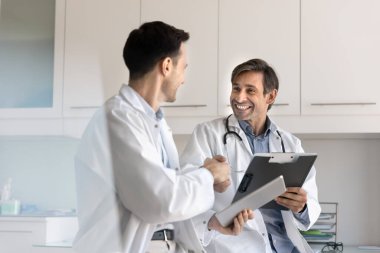 Profesyonel işbirliği, anlaşma, tıbbi alanda karşılıklı saygı. Beyaz önlüklü, gülümseyen iki Latin erkek doktor el sıkışıyor klinikte olumlu ve başarılı bir etkileşim içindeler.