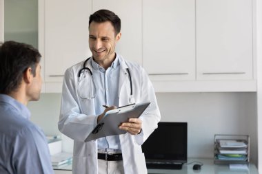 Üniformalı orta yaşlı doktor bilgi yazıyor, not alıyor, tıbbi kayıtları güncelliyor, veya klinikteki hastaya danışmanlık yaparken reçete yazıyor, gülümsüyor, pozitif etkileşime giriyor.