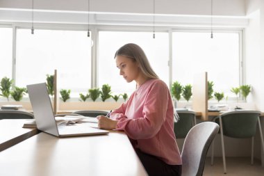 Dijital eğitim. Kampüs kütüphanesinden uzak öğrenim gören genç kız liseli öğretmenlerin sanal konferans videolarını dinleyip not defterinden notlar alıyorlar.
