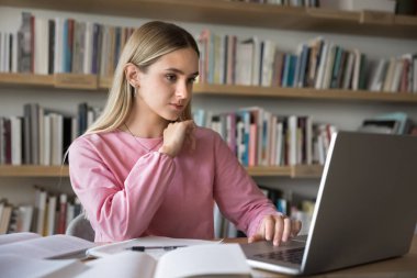 Bilgi aramak için eğiliyorum. Konsantre olmuş milenyum öğrencisi kız çeşitli eğitim materyali kaynaklarıyla çalışıyor. Bilgisayar üzerinde bilimsel web siteleri okuyup, el yazısı notlar alıyor.