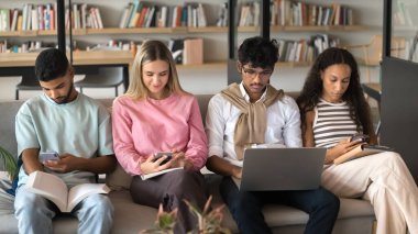 Modern çalışma oturumu. Çeşitli etnik çeşitlilikteki genç öğrenciler, kampüs kütüphanesinde koltuklarda otururlar. Kişisel eğitim görevlerinde kendi kendini eğitmeyi öğrenmek için kağıt parçaları kullanırlar.