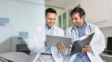 Klinikte dijital tablet kullanan iki gülümseyen erkek doktor, teletıp platformu ya da uygulaması aracılığıyla iş arkadaşlarıyla ya da uzmanlarla iletişim kuruyor ya da yenilikçi yapay zeka teknolojilerini gözden geçiriyorlar.