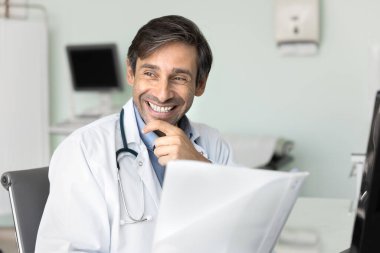 Mutlu gülüşü ve düşünceli ifadesiyle orta yaşlı, profesyonel doktor masasına oturur, kağıtları tutar, başarılı tıbbi sonuçları gözden geçirir, etkili bir tedavi planı bulur.