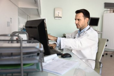 Bilgisayarda çalışan odaklanmış adam doktor ofiste oturur, bilgisayar ekranını gösterir, hasta hastalıkları hakkında önemli bilgiler öğrenir, tıbbi kayıtları inceler, tanı veya tedavi planını inceler.