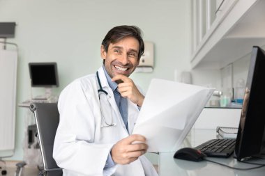 Mutlu adam kardiyolog, beyaz üniformalı terapist çağdaş danışma odasında masasında oturuyor, iş akışından ve evrak işlerinden dikkati dağılmış, mutlu bir gülümsemeyle mesafeye bakıyor. Görev.