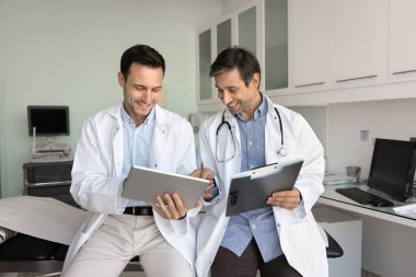 Tablet ve not panosundaki bilgileri incelerken gülümseyen iki Latin doktor, profesyonel bir tartışma içindeler, hasta verilerini, tıbbi kayıtlarını veya araştırmalarını inceliyorlar. Profesyonellik, yenilik