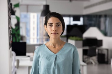 Ofiste portre için poz veren kendine güvenen genç Latin iş kadını, kameraya bakıyor, modern ofis alanında tek başına duruyor. İş projesi lideri, şirket yöneticisi portresi