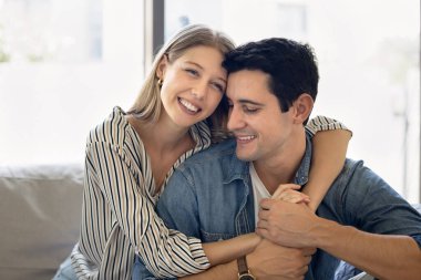 Sevecen Arjantinli kadın, sevgili İspanyol erkeğine sarılıyor, gülüyor, günışığıyla dolu modern oturma odasında rahat bir kanepede birlikte oturuyor. Kiracılar ya da ev sahipleri genç aile portresi