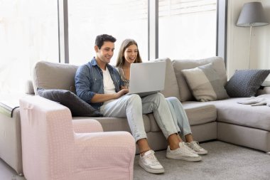 Latin çift, rahat oturma odasındaki rahat modern koltukta dizüstü bilgisayarlarıyla oturup rahatlama ve internet kullanımının tadını çıkarıyorlar. Seyahat rezervasyonu, uçak ya da tatil kiralama, sipariş verme, mal alma.