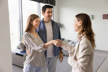 Mutlu çift, kadın emlakçıyla el sıkışıyor, modern bir dairede duruyor, kiralıyor, anlaşmadan memnun kalıyorlar. Gayrimenkul işlemleri, ev sahipliği ve profesyonel danışmanlık