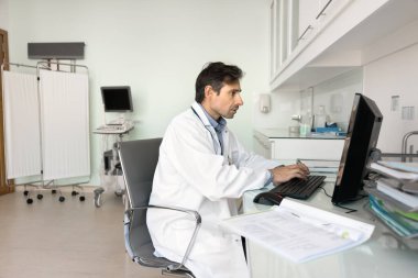 Orta yaşlı erkek doktor masa başında oturur, hasta kayıtlarını inceler, sağlık geçmişini ve test sonuçlarını öğrenir, önemli notlar ekler, metinleri analiz eder, tedavi planını düşünür, ilaç yazmayı düşünür.