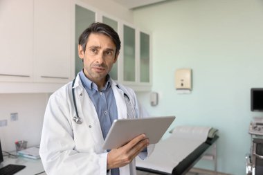 Düşüncelere dalmış düşünceli pratisyen hekim, hasta durumundaki tedavileri, ilaçları ya da terapiyi göz önünde bulundurarak muayene odasında duruyor, semptomları değerlendiriyor, kararlar veriyor, dijital tablet üzerinde çalışıyor.