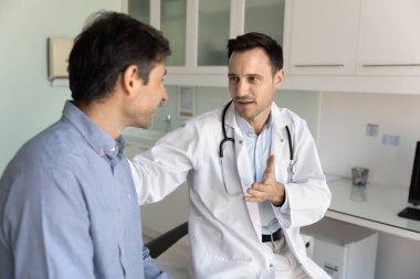 Beyaz önlüklü orta yaşlı profesyonel erkek doktor hastanın omzuna dokunuyor, rahatlama sağlarken dostça tavır sergiliyor, tedavi planını açıklıyor, klinikteki teşhisten bahsediyor