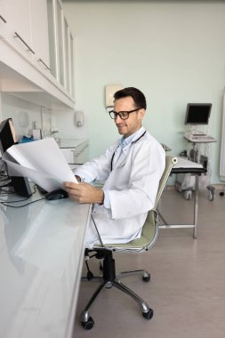 İş yerinde çalışan üniformalı doktor evrakları tutuyor, hasta kayıtlarını inceliyor, tıbbi geçmişi veya test sonuçlarını analiz ediyor, taramaları yorumluyor, tedavi planlarını hazırlıyor, ilaçlara veya terapiye karar veriyor.