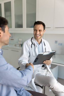 Beyaz üniformalı pozitif İspanyol erkek doktor hasta ile el sıkışıyor, güven, anlaşma ve tıbbi randevu sırasında pozitif iletişim gösteriyor. Muayenenin sonu, güven