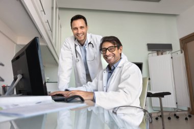 Danışma odasında birlikte çalışan iki orta yaşlı erkek kardiyoloğun portresi, neşeli bir ifadeyle kameraya bakıyor, profesyonelliği, güveni ve yeterliliği yayıyor. Sağlık hizmetleri