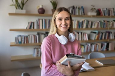 Modern zaman öğrencisiyim. Portre mutlu genç kız lise öğrencisi kampüsteki kitaplıklarda poz veriyor ya da kütüphanede uzaktan öğrenim için kulaklıkla kağıt çalışma kitaplarına bakarak.