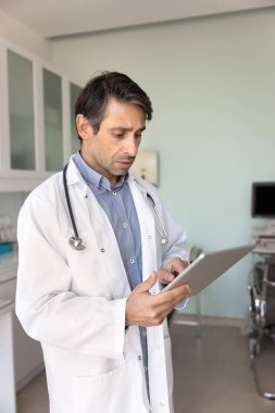 Odaklanmış doktor, dijital tablet kullanarak, klinikte ayakta durarak, anestezi veya geçmişini kontrol ederek, raporlar yazarak, bilgileri güncelleyerek,