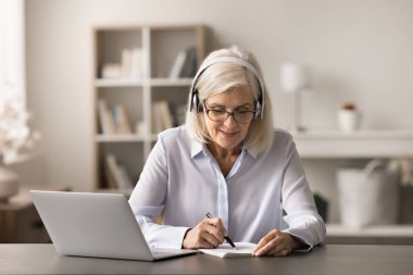 Sanal eğitim ve profesyonel beceri gelişimi. Kulaklıklı, konsantre yaştaki iş kadını ofis dizüstü bilgisayarındaki video sunumu atölyesi atölyesindeki el yazısı notlarını izleyip internetten öğreniyor.