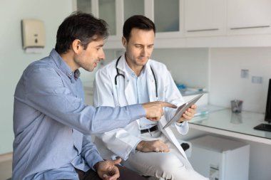 Kardiyolog, özel klinik muayenehanesine yaptığı ziyarette, beyaz önlüklü danışman hasta kılığında, dijital tabletin kalp muayenesi sonuçlarını inceliyor. Modern teknoloji, profesyonel sağlık hizmetleri