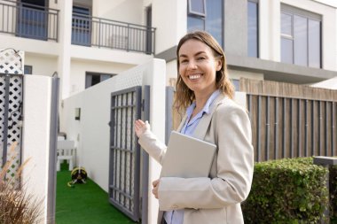 Zarif bir ceket giymiş güzel bir emlakçı kameraya gülümseyerek bakıyor, davetkar bir şekilde el kol hareketi yapıyor, müşterileri modern konutlara kabul ediyor, satılık ya da kiralık ev sunuyor.