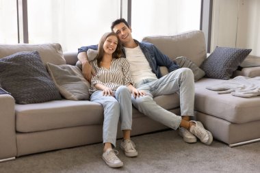 Modern oturma odasındaki rahat koltukta oturan güzel Latin çift kameraya gülümseyerek bakıyor, kiralık dairede rahatlığın tadını çıkarıyor. Ev sahipleri, yeni evlilerin ev portresi. Banka kredisi, yeni ev