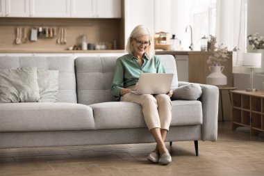 Laptop ve emeklilik. Modern Ev İçi 'nde rahat bir kanepede dizüstü bilgisayarda çalışan mutlu yaşlı bayan hobi eğlence parşömeni sosyal medya sayfaları için internette serbest iş arama bilgileriyle meşgul.