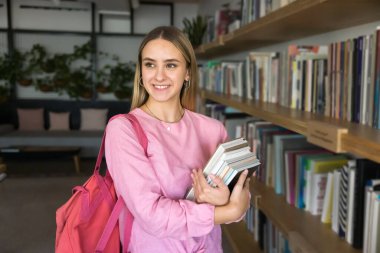 İyi eğitim parlak perspektifleri açar. Gülümseyen gen z kız öğrenci kütüphanede duruyor kitap yığını kenara bakın akademik araştırmalar için akademik diploma almak için profesyonel edebiyat bulduğu için mutlu hissedin