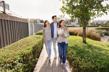 Emlakçı genç çiftlere yerleşim bölgelerinde rehberlik ediyor, yeni ev ve toplum özelliklerini sergiliyor, yeşil ağaçlarla korunan patika boyunca yürüyor, güneşli yaz günlerinde çalılar arasında yürüyor.