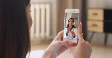 Kadın eller akıllı telefonu, doktor görüntüsü cihaz ekranında, omuz üstünden görüntüyü kapatıyor. Uzaktan kumandalı tıbbi danışmanlık için ilaç platformunu kullanarak teletıp