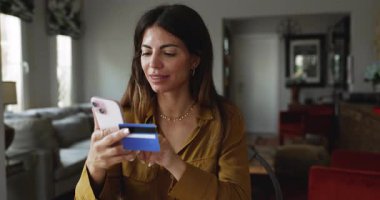 Gülümseyen Latin kadın elinde kredi kartı ve akıllı telefon tutuyor, çevrimiçi alım yapıyor, ödeme bilgilerini uygulamaya geçiriyor, banka bakiyesini veya kart detaylarını kontrol ediyor, ödeme faturasını ödüyor, elektronik servislere abone oluyor.