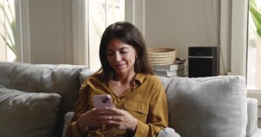 Genç Latin bir kadının modern akıllı telefonuyla evdeki koltuğunda oturması, mobil uygulama veya sosyal ağlar aracılığıyla arkadaşlarıyla veya ailesiyle sohbet etmesi, dijital iletişimin ve yeni uygulama kullanımının keyfini çıkarması