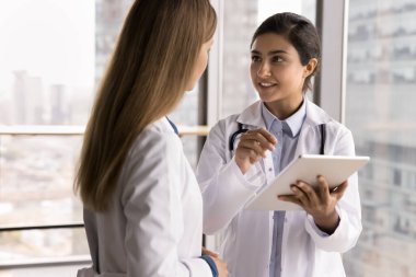 Sorunları birlikte çözmek. Genç kadın doktor tıp çalışanı kadın eczacı hekimle konuşup tablet bilgisayar ekranındaki sağlık araştırması makalesini tartışır, profesyonel görüş alır.