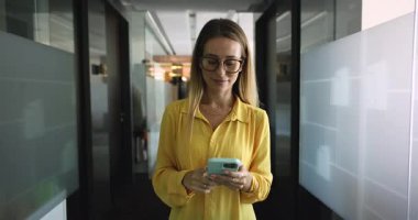 İş yerinde çalışan genç bir kadının iş arasında cep telefonuyla yazı yazması, ofis koridorunda kameraya yürümesi, gülümsemesi, yüzünü çevirmesi, çevrimiçi iletişimi düşünmesi.