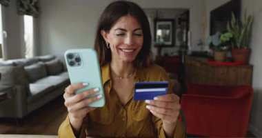 Çekici İspanyol bir kadın evde online alışveriş yapıyor, akıllı telefon kullanıyor, kart satın alıyor, inceleme maliyetleri şaşırtıcı ya da endişe verici, cep telefonu uygulaması üzerinden para transfer ediyor.